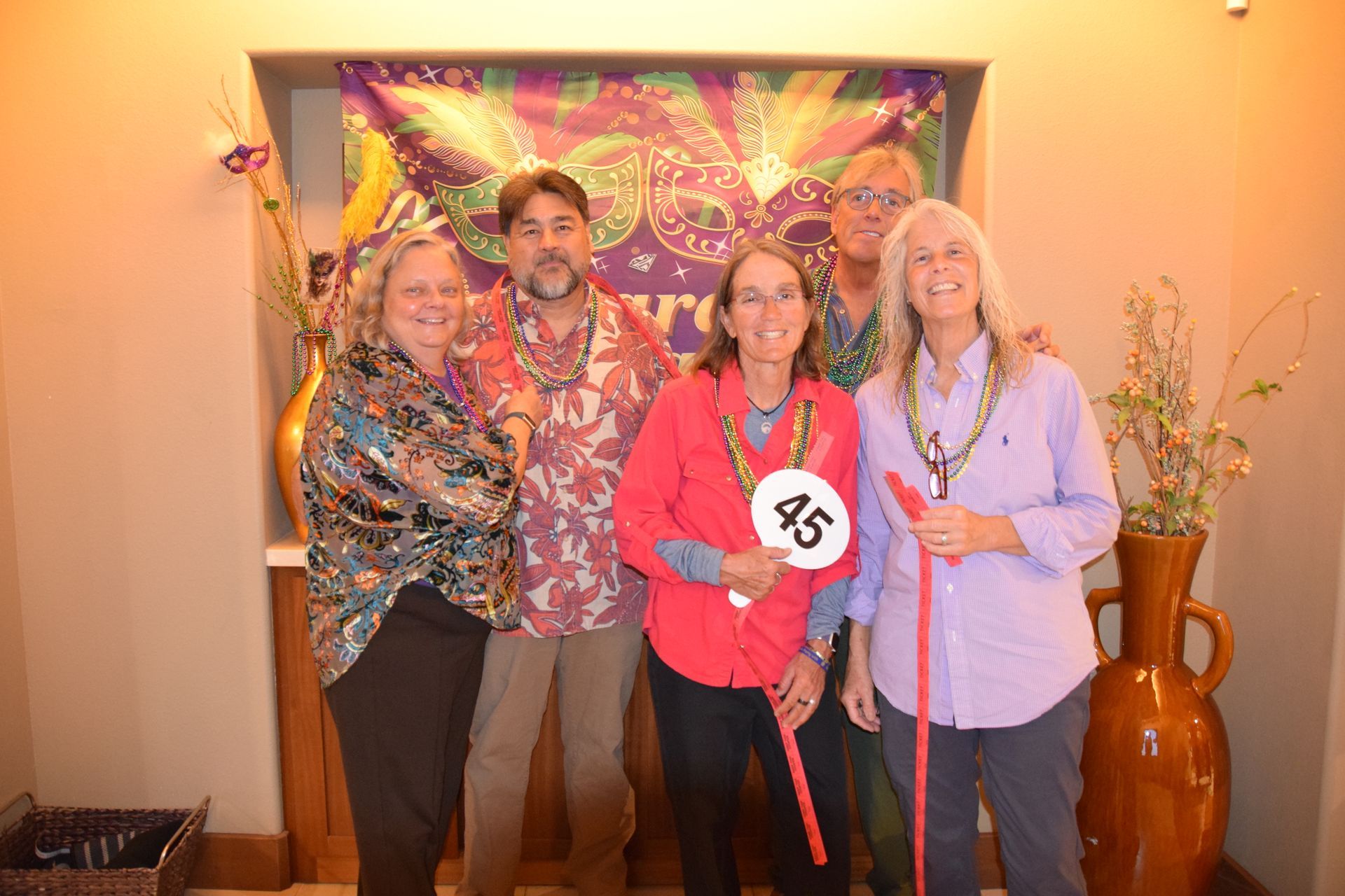 Four people standing before a decorative Mardi Gras backdrop; the person second from right holds a sign with the number 45.