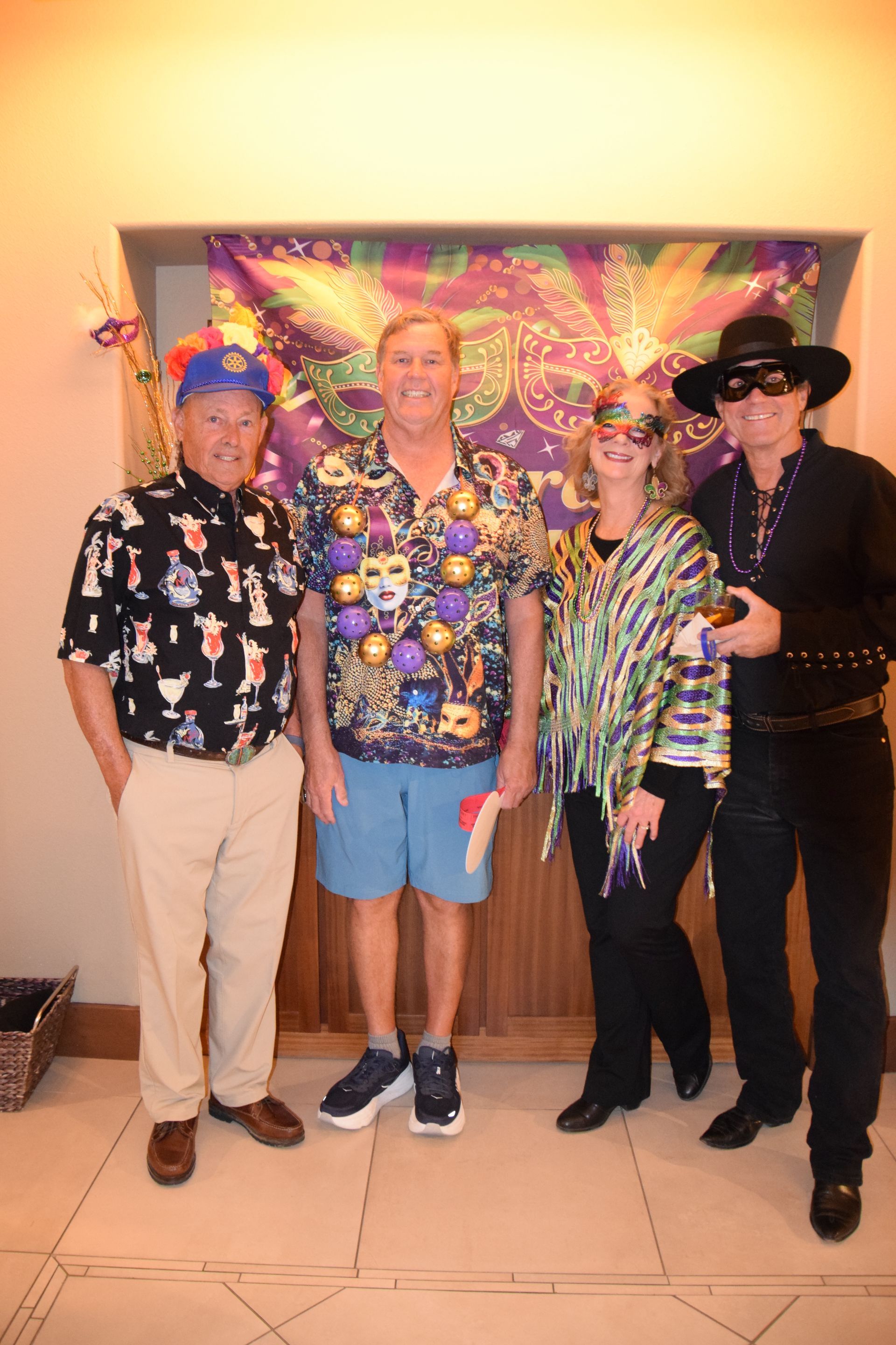 Four people in festive attire stand before a Mardi Gras-themed backdrop at an indoor event.