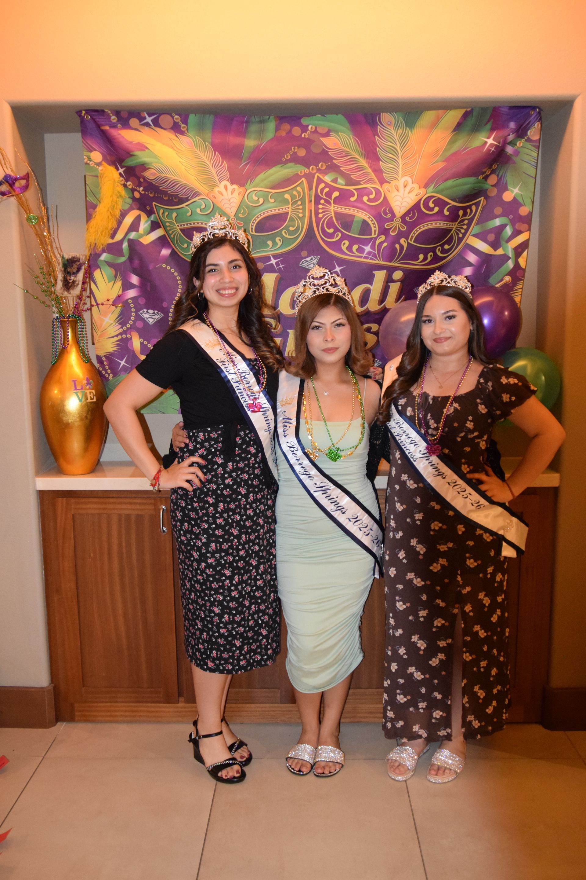 Three people wearing crowns and sashes stand posing in front of a Mardi Gras themed backdrop.