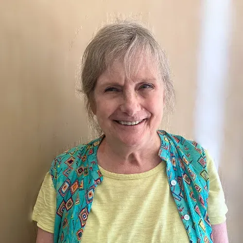 A smiling person with shoulder-length hair wearing a yellow shirt under an unbuttoned turquoise patterned vest.