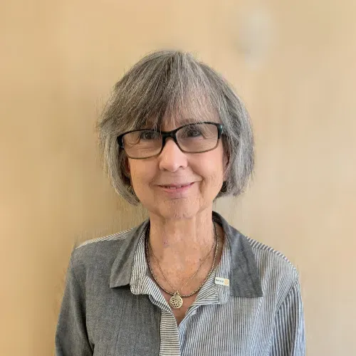 A person with grey hair and glasses smiling against a solid tan background, wearing a grey striped, button-down shirt.