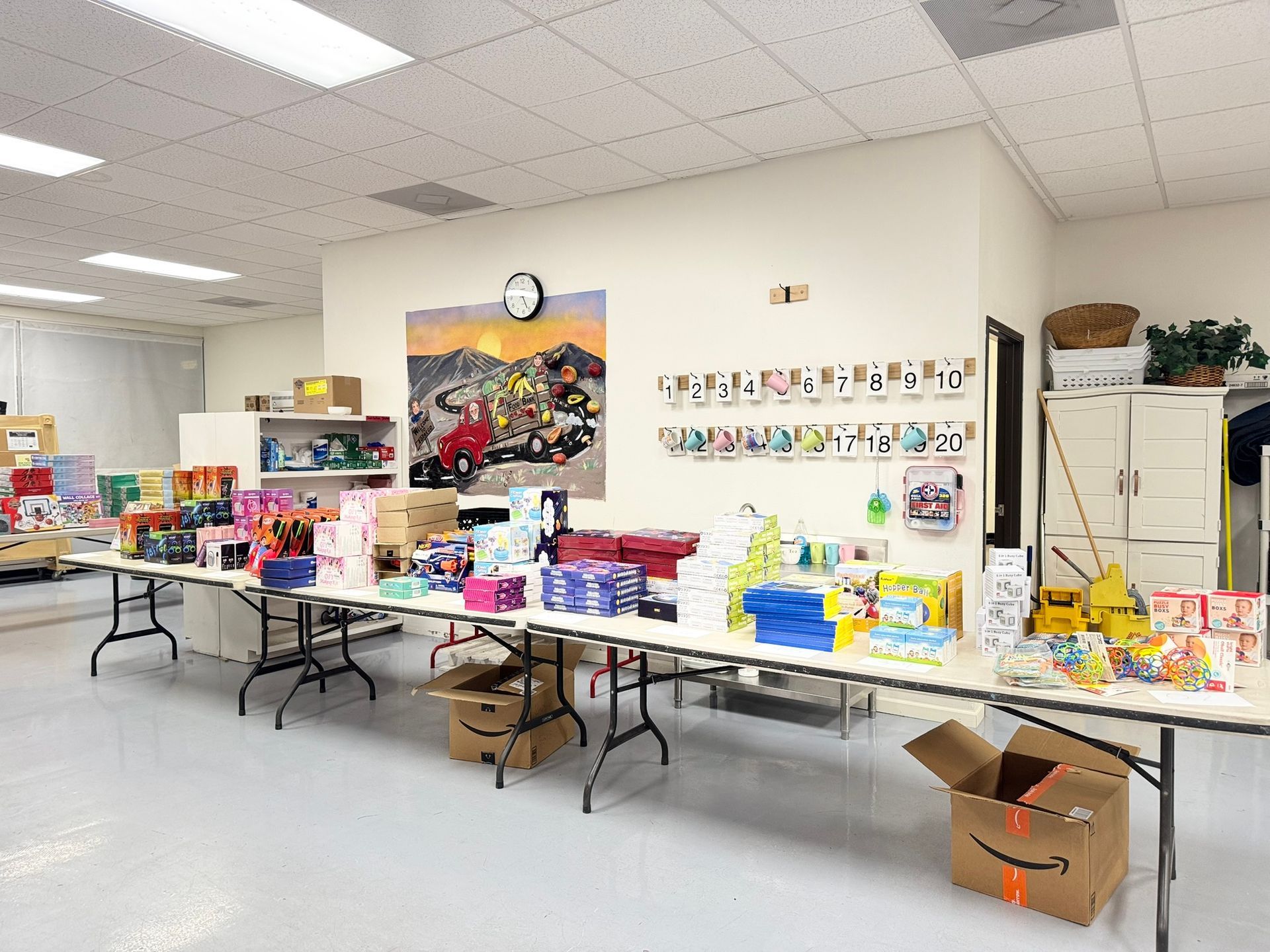 Long folding tables in a room are covered with various supplies, including boxes, papers, and books for distribution.