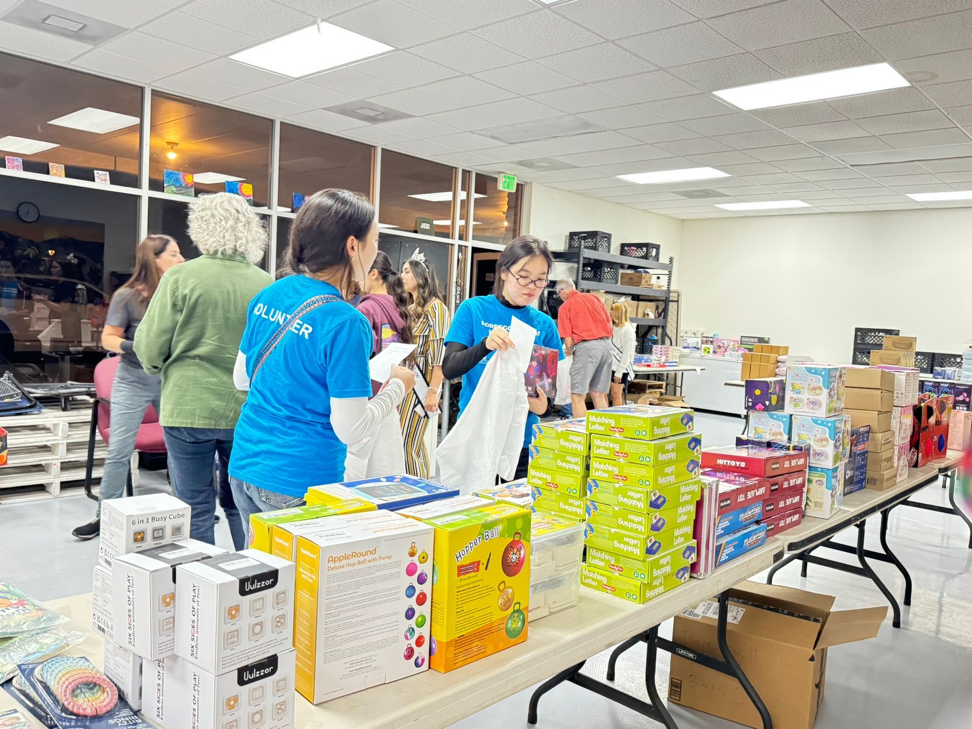 Volunteers in blue shirts sort and pack supplies on tables inside a community center or warehouse.