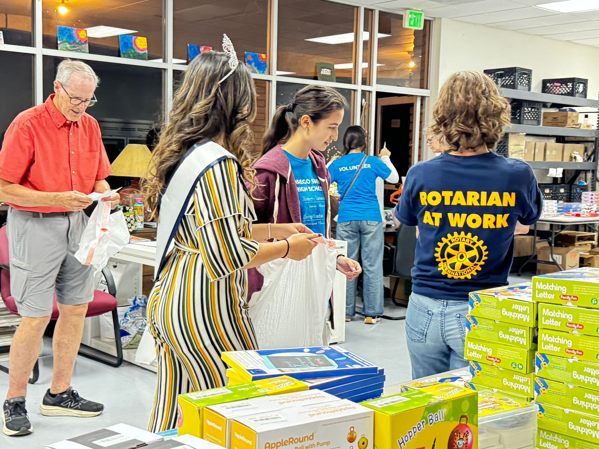 Volunteers pack boxes of supplies in a warehouse setting, with one person wearing a shirt that says 