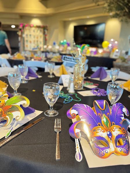 A Mardi Gras-themed table setting with decorative masks, beads, purple napkins, and glassware on a black tablecloth.