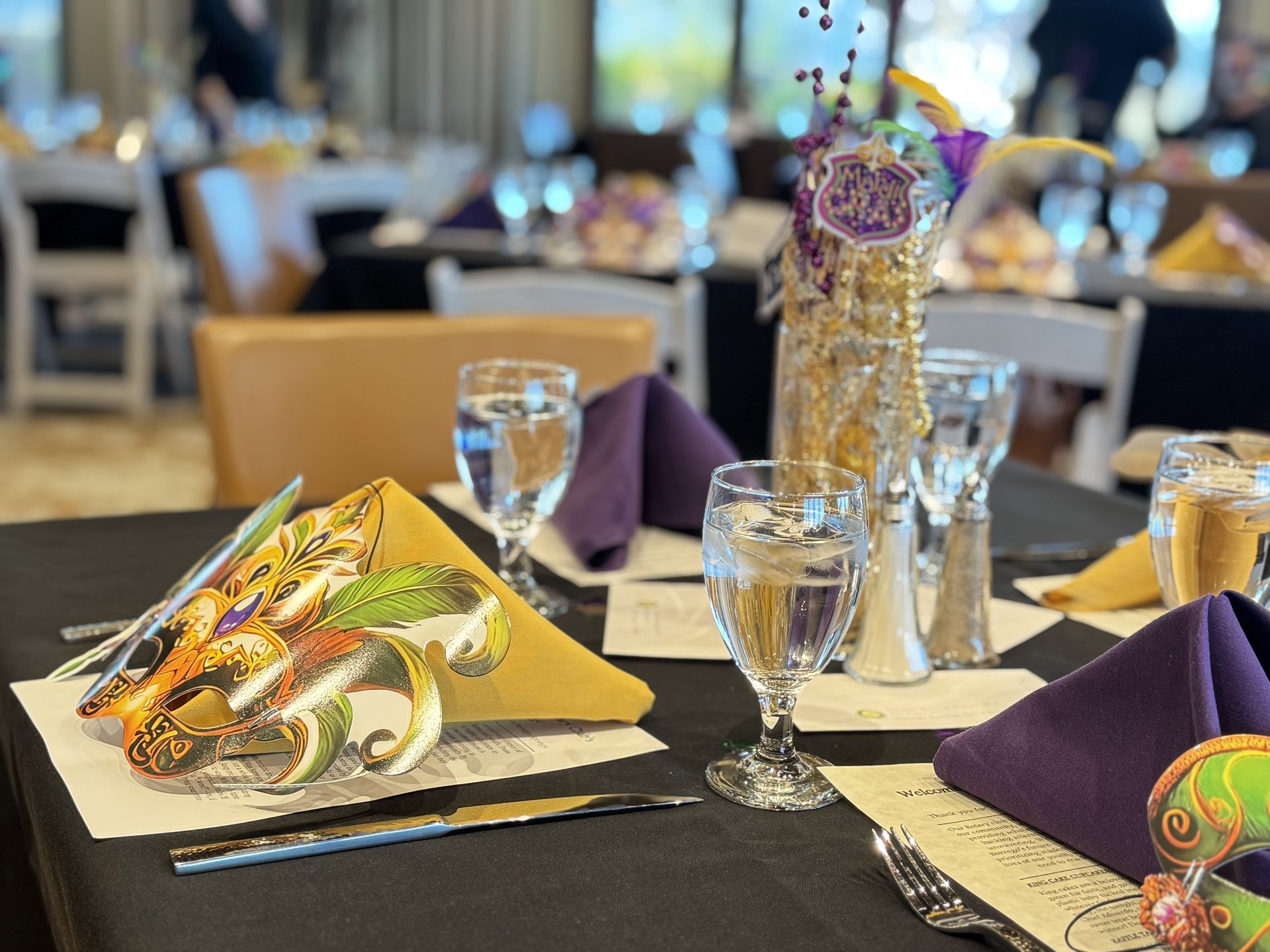 A table set for a Mardi Gras-themed event with purple napkins, gold decorations, and glasses on a black tablecloth.