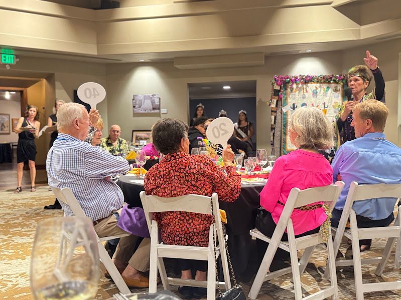 People seated at a table during an auction, holding up numbered paddles with one person gesturing in a bright room.