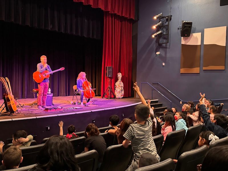 A musician playing guitar and a cellist perform on a theater stage before an audience of children with their hands raised.