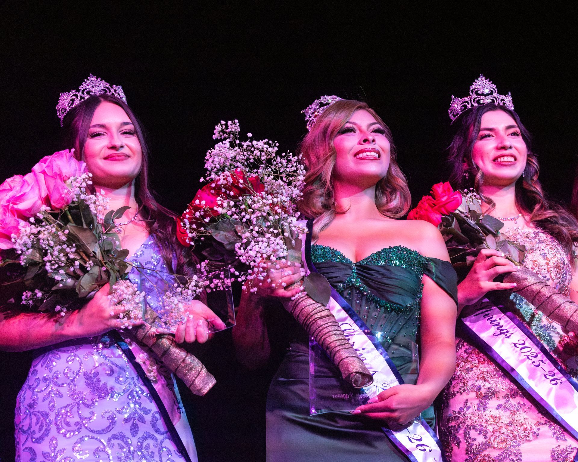 Three people in evening gowns and crowns hold bouquets of flowers on a stage.