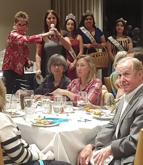 A woman speaks into a microphone while others wearing sashes and crowns stand behind seated guests at a formal event.