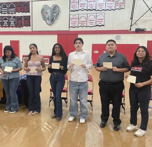 Six people stand in a line on a gymnasium floor, each holding a small card while facing the camera.