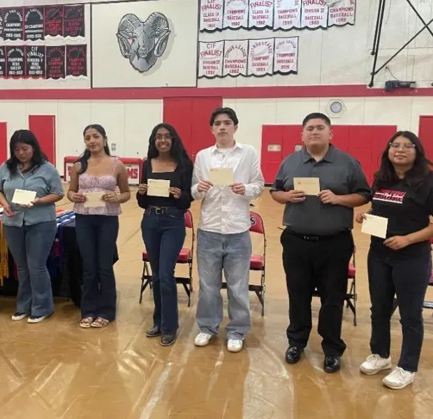 Six people stand in a line on a gymnasium floor, each holding a small card or certificate.