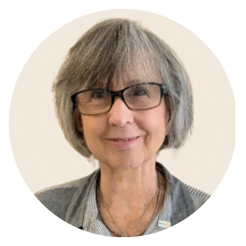 A person with grey hair and glasses smiling against a solid tan background, wearing a grey striped, button-down shirt.