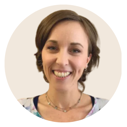A smiling person with brown hair, wearing a floral print top and a double-layered necklace, stands against a beige wall.