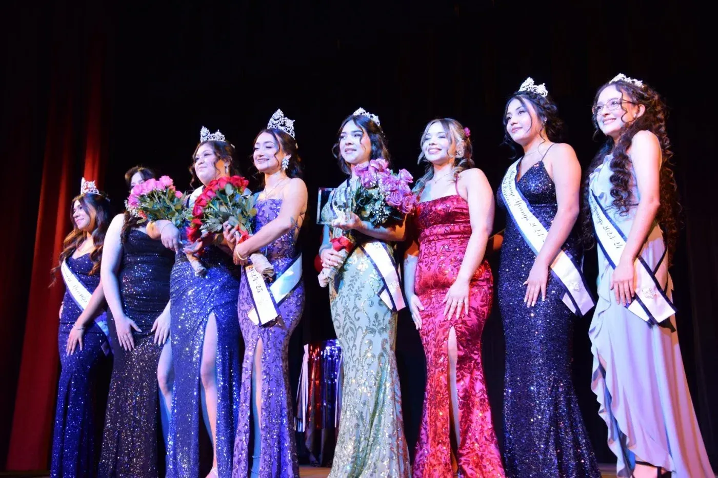 Eight contestants in formal gowns and sashes pose with bouquets on a stage with a dark red curtain backdrop.