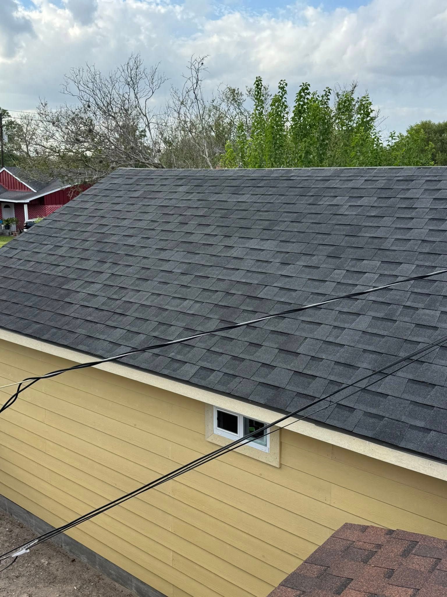 Gray shingled roof and beige siding with a window, viewed from above with trees in the background