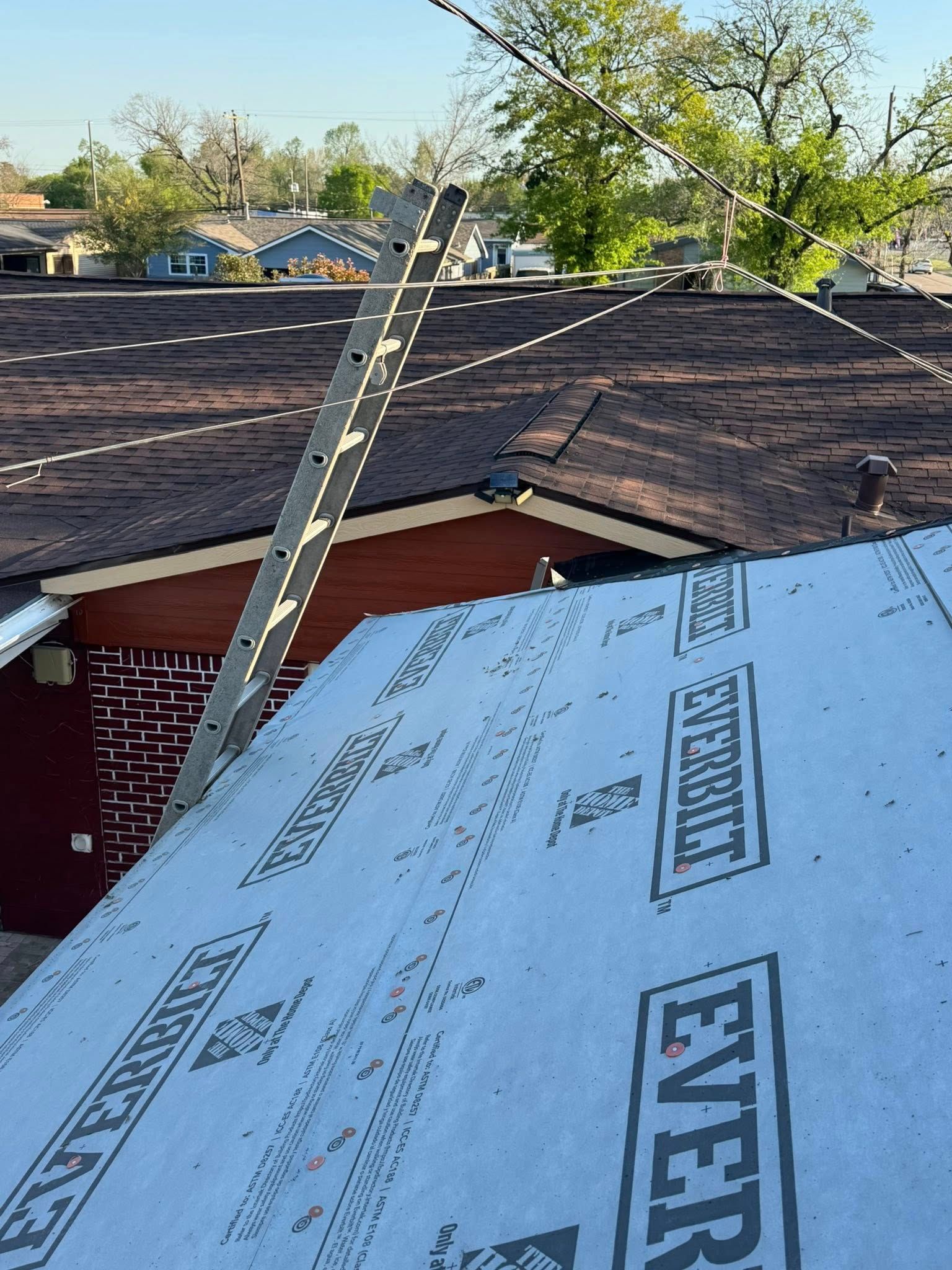 Roofing underlayment on a sloped roof beside a ladder, with neighboring houses and trees in the background