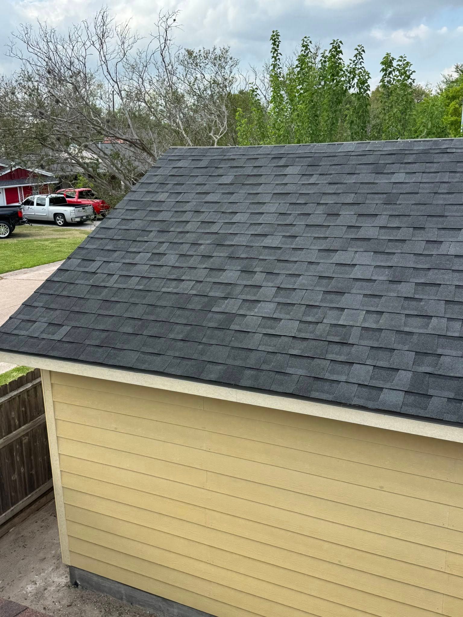 Yellow shed with a dark shingled roof in a backyard, with trees and a red truck in the background