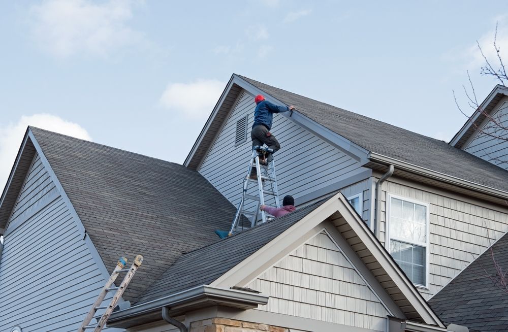 Workers on a rooftop repairing gray shingles on a house with a ladder.