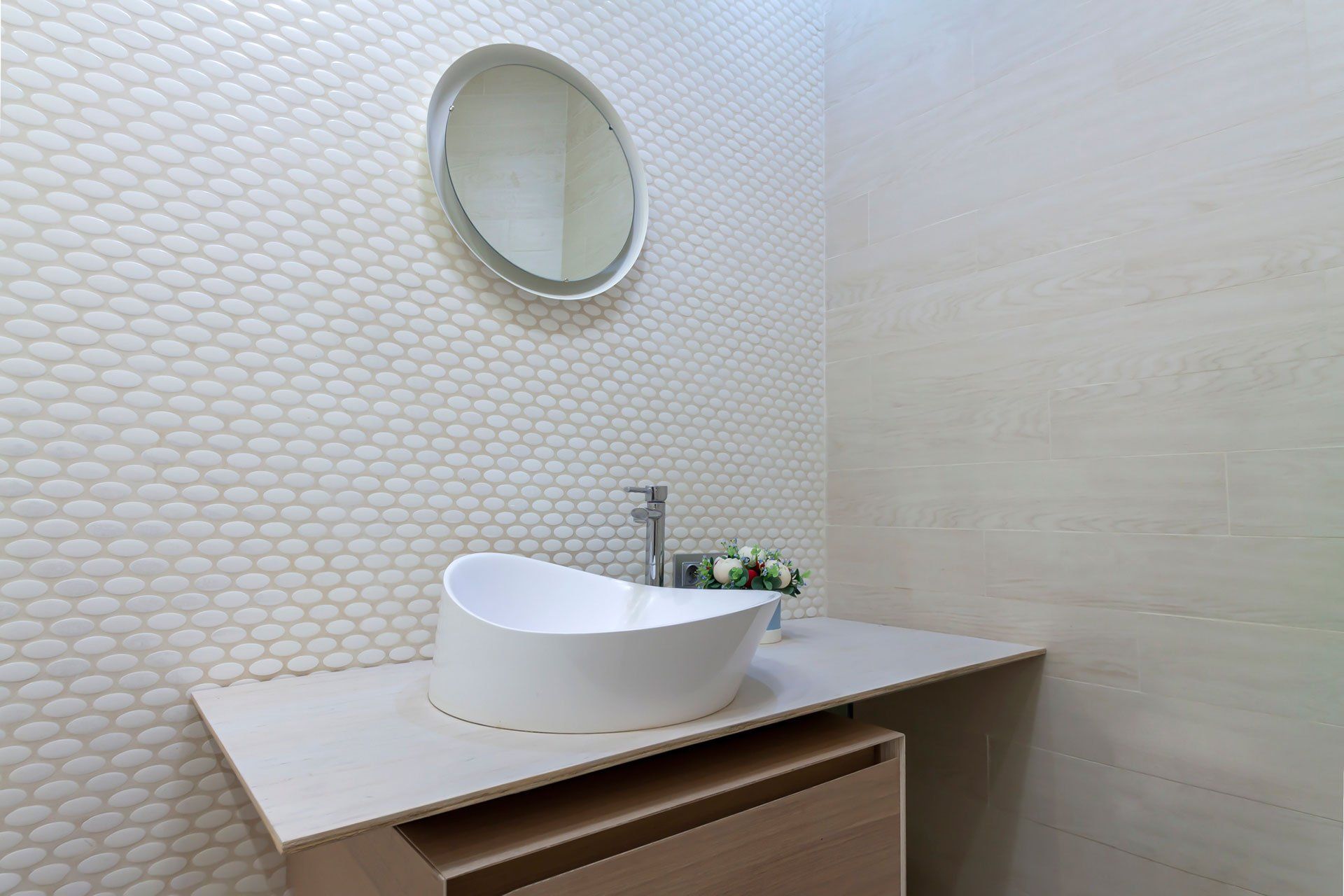 White washbasin with faucet on wooden countertop in minimalist modern bathroom. Scandinavian interior with tiled mosaic wall wall and round mirror. Copy space and nobody.
