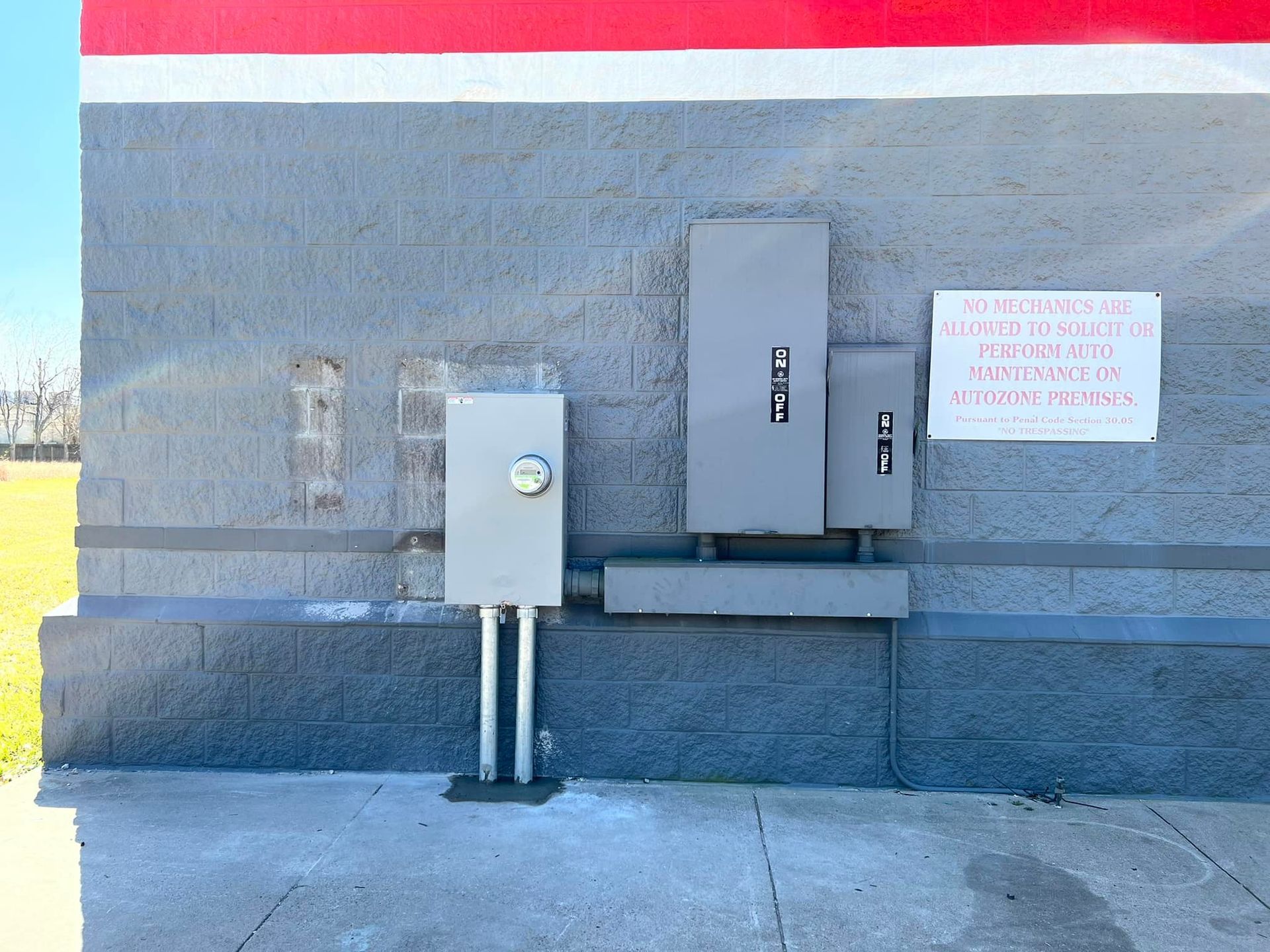 Electrical boxes on a gray brick wall with a red and white stripe above. A sign is to the right.