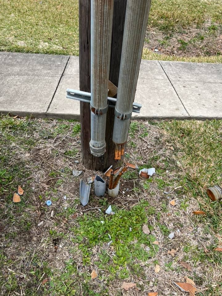 A damaged utility pole with exposed wires at its base, set in a grassy area near a sidewalk.