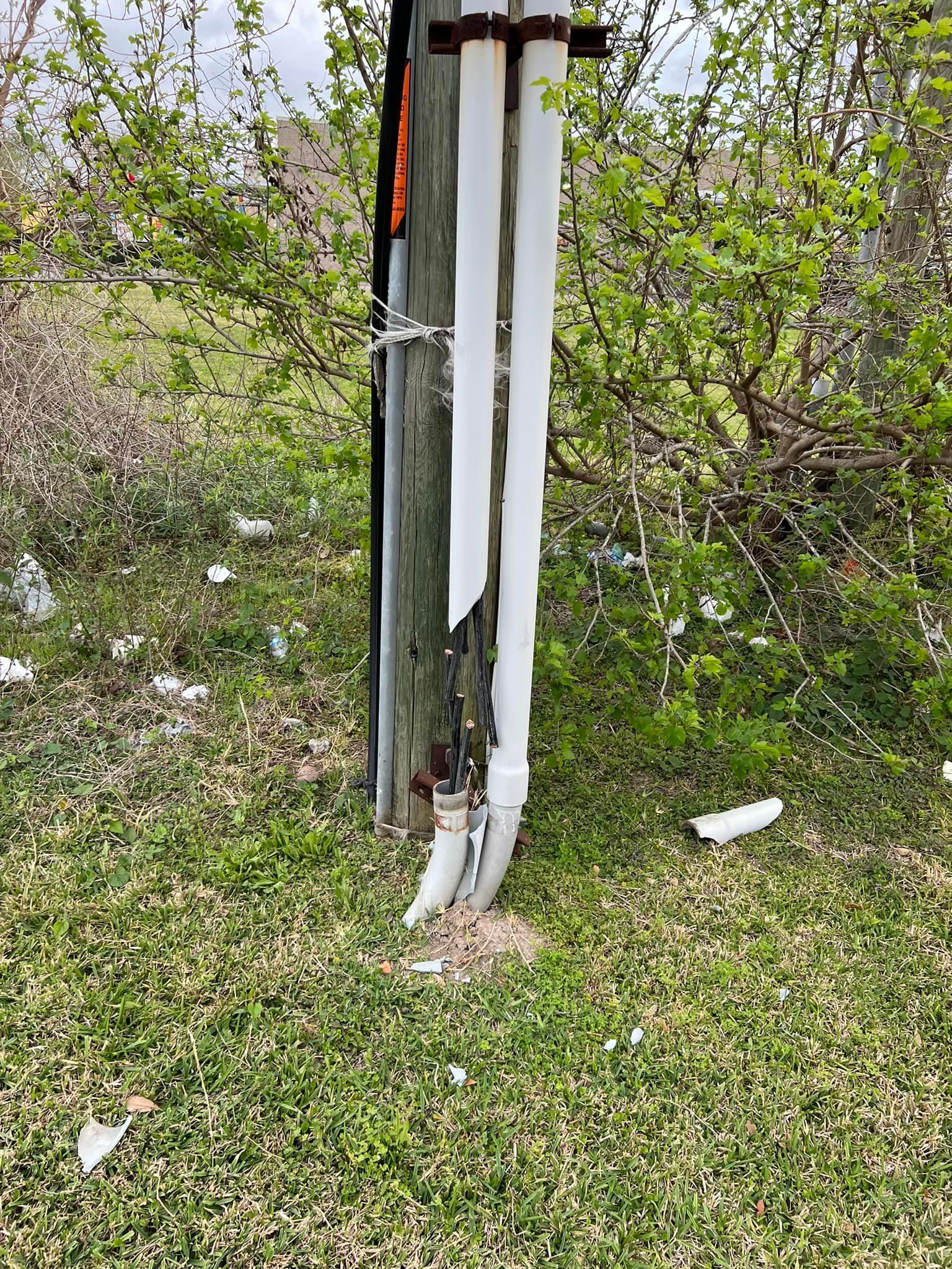 Utility pole with damaged white covering, wires exposed, near green grass and bushes.