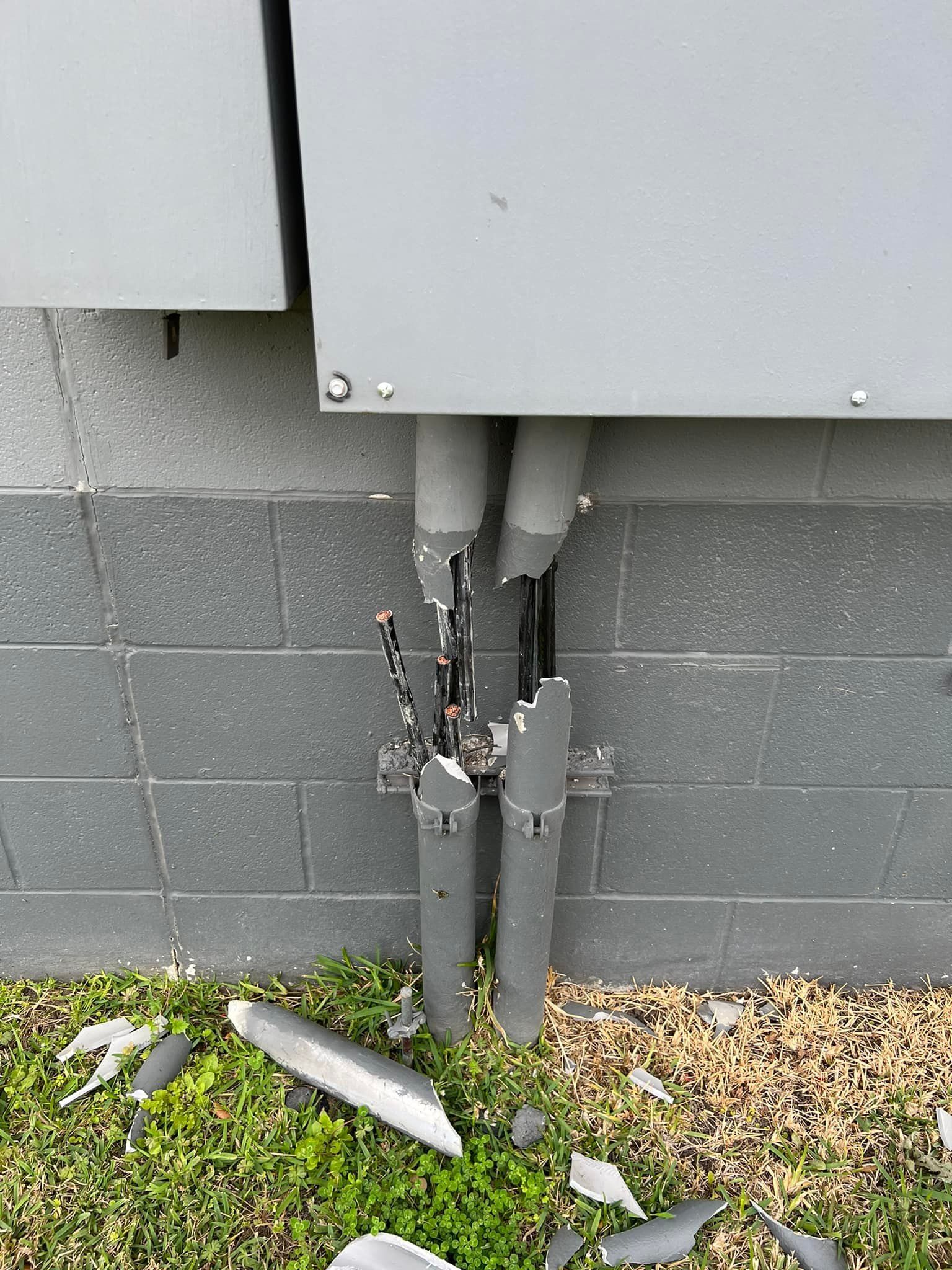 Damaged electrical conduit and wires protruding from a gray brick wall, with broken pieces on the grass.