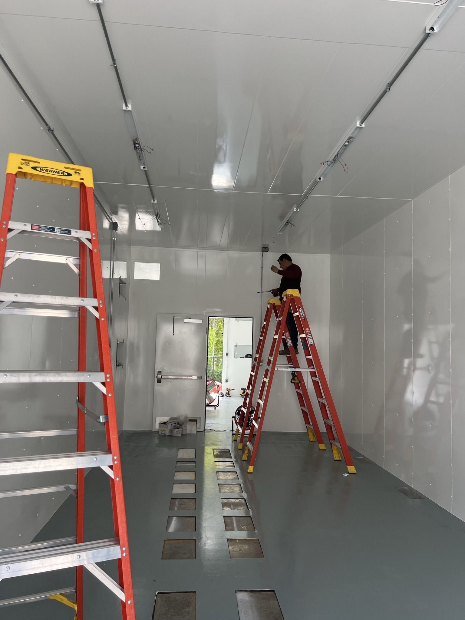 Person on a ladder installs ceiling fixtures in a white-walled room with a gray floor.