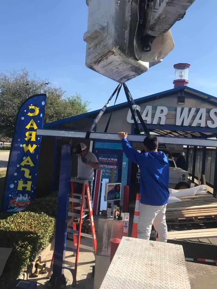 Two men lift a large car wash sign with straps, near a car wash building, blue sky.