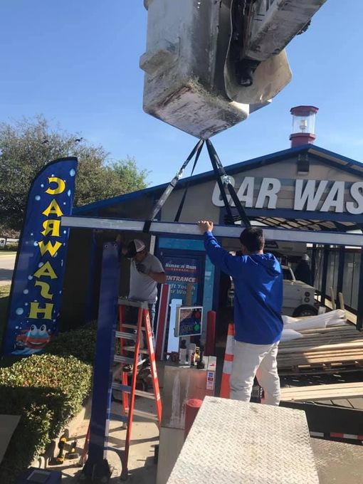 Two men lift a large car wash sign with straps, near a car wash building, blue sky.