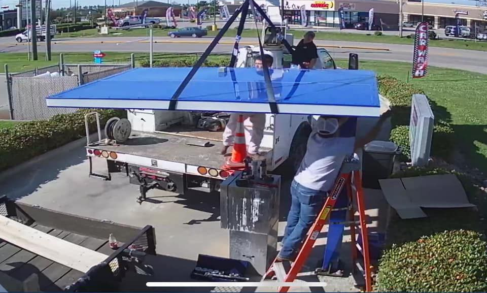Workers installing a large blue sign on a truck bed with a crane, outdoors on a sunny day.