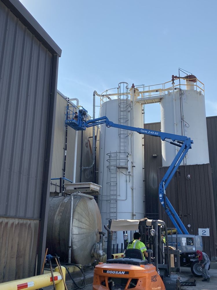 Blue lift reaching toward industrial tanks next to a building. A worker in a yellow vest and a forklift are at the base.