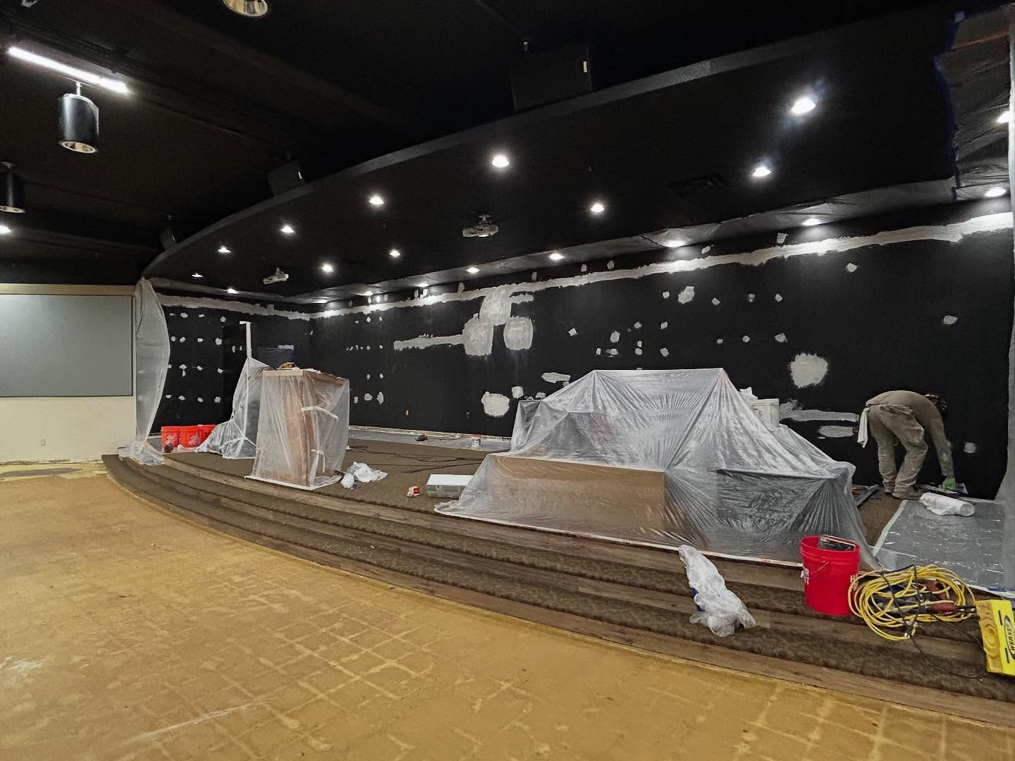 Construction in a black-walled room with a stage. Covered equipment, a worker, and exposed drywall are visible.