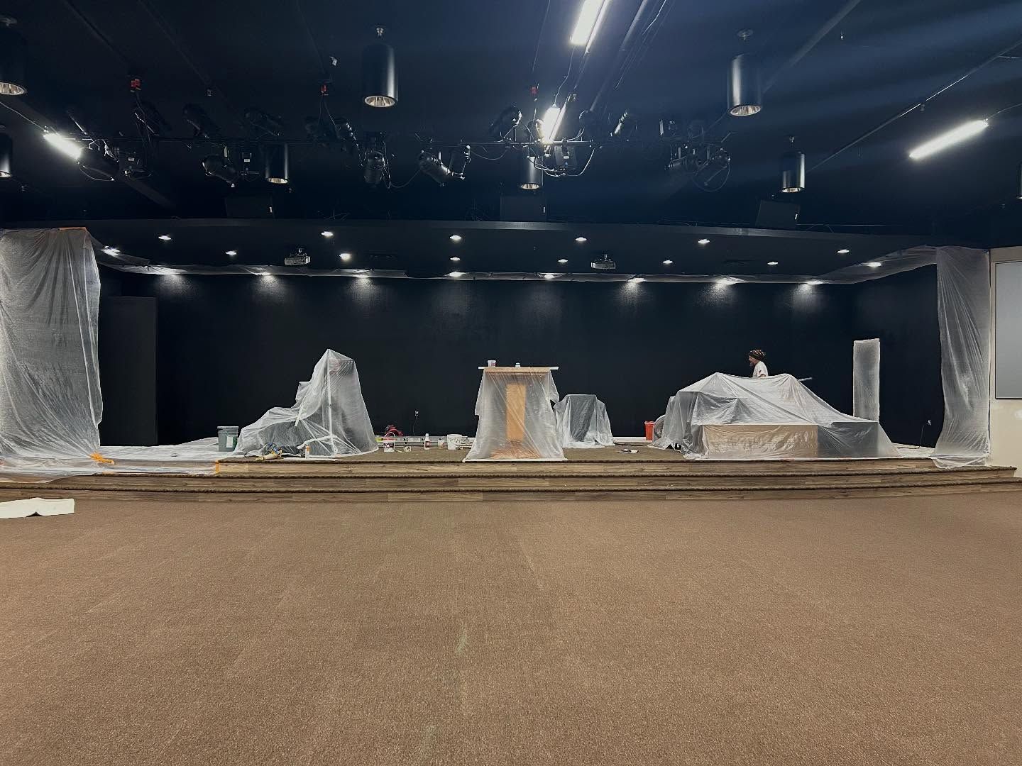 A church stage under renovation. The stage is covered in plastic. Black backdrop. Brown carpeted floor.