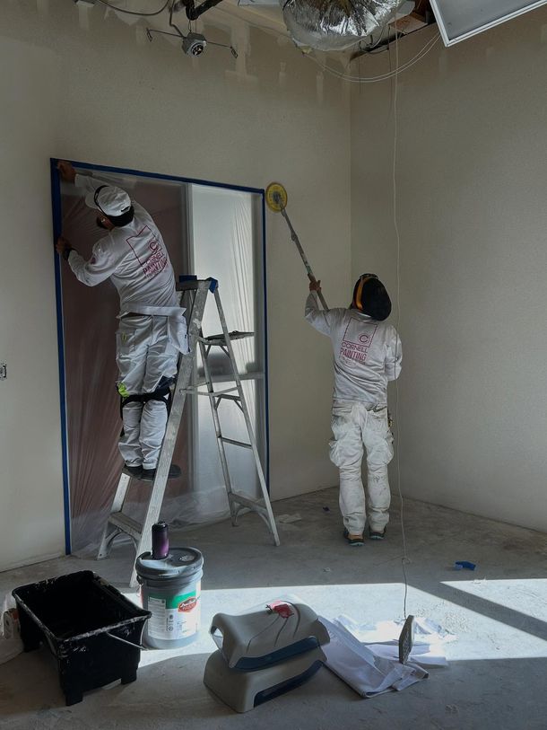 Two painters in white suits painting a room; one on a ladder taping a door frame, the other using a roller.