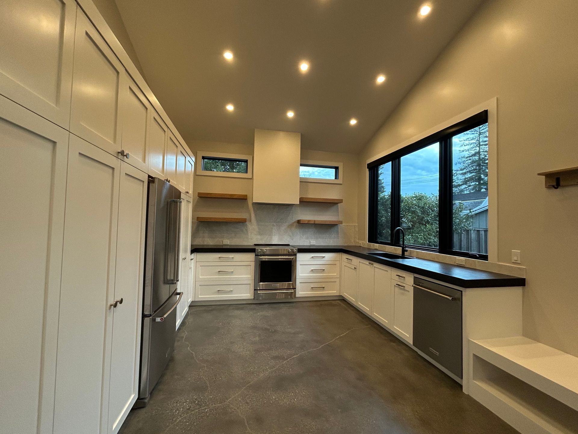 Modern white kitchen with stainless steel appliances, black countertops, and concrete floor.