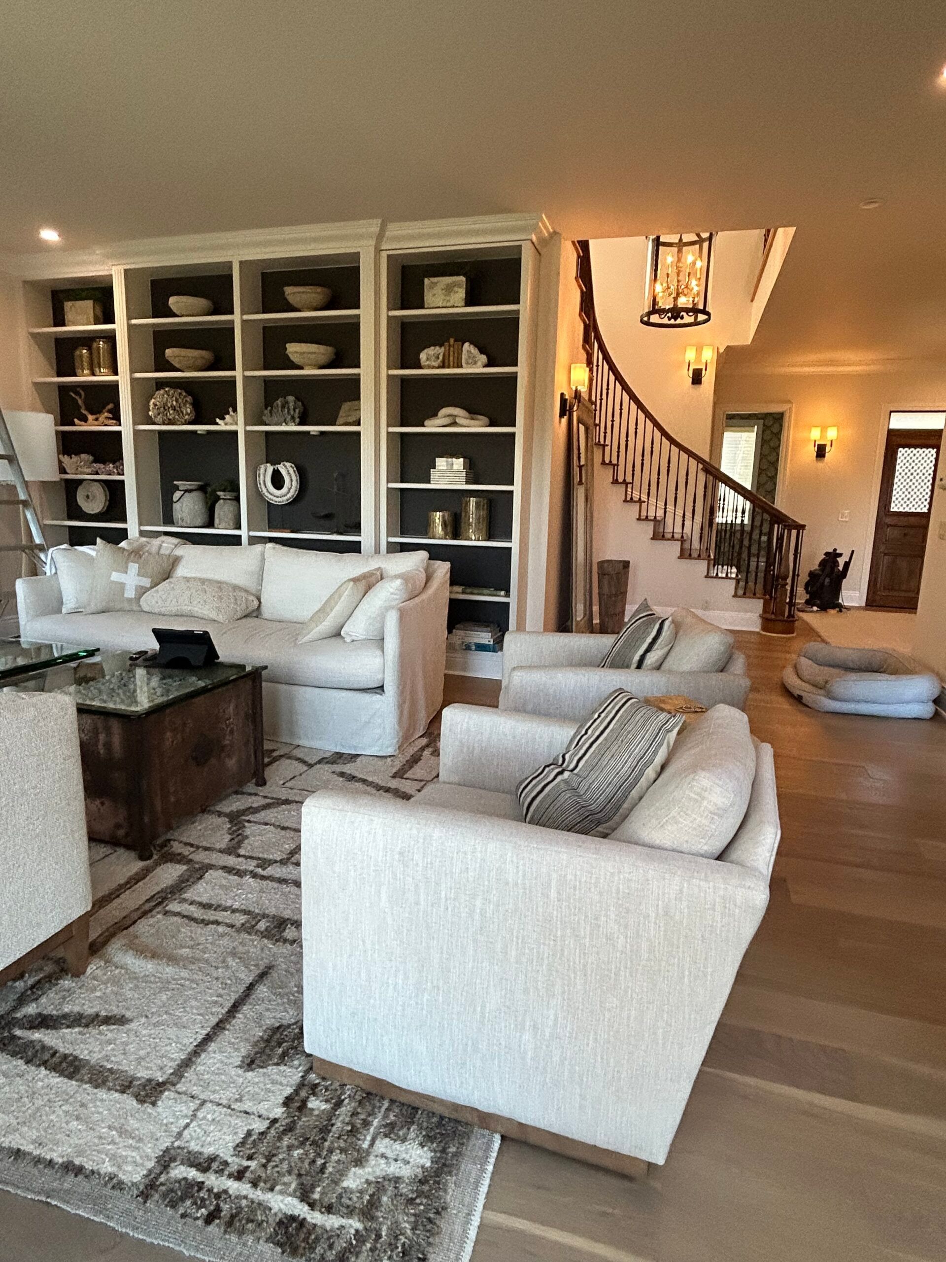 Living room with white bookshelves, couches, chairs, and a staircase in a house.