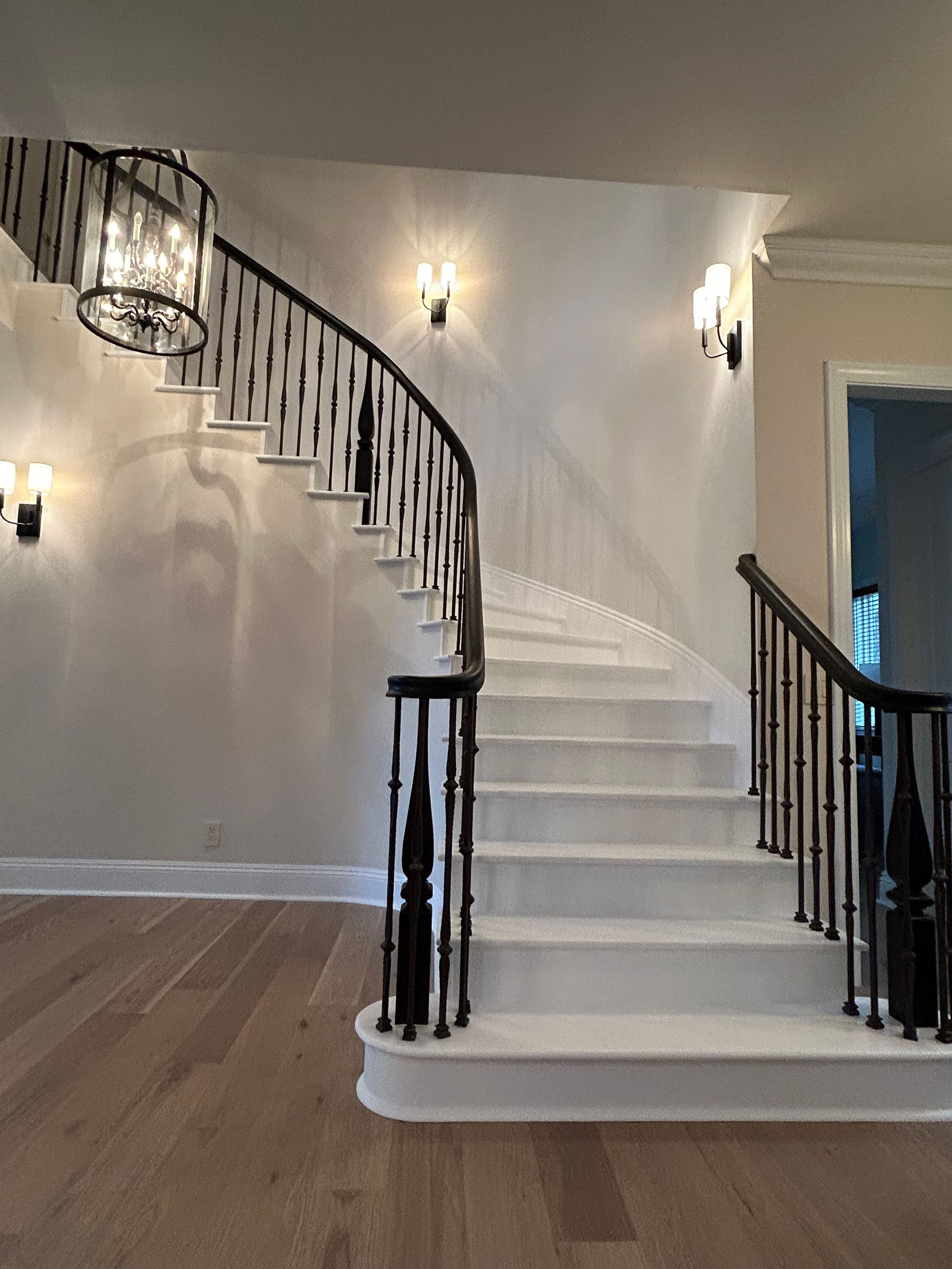 Staircase with black railing and white steps, illuminated by wall sconces and a chandelier. Hardwood flooring.
