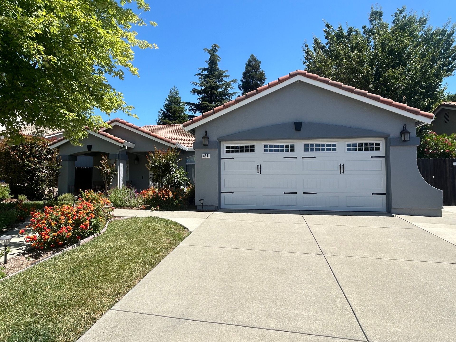 Gray house with white garage door, red tile roof, concrete driveway, and green lawn under blue sky.