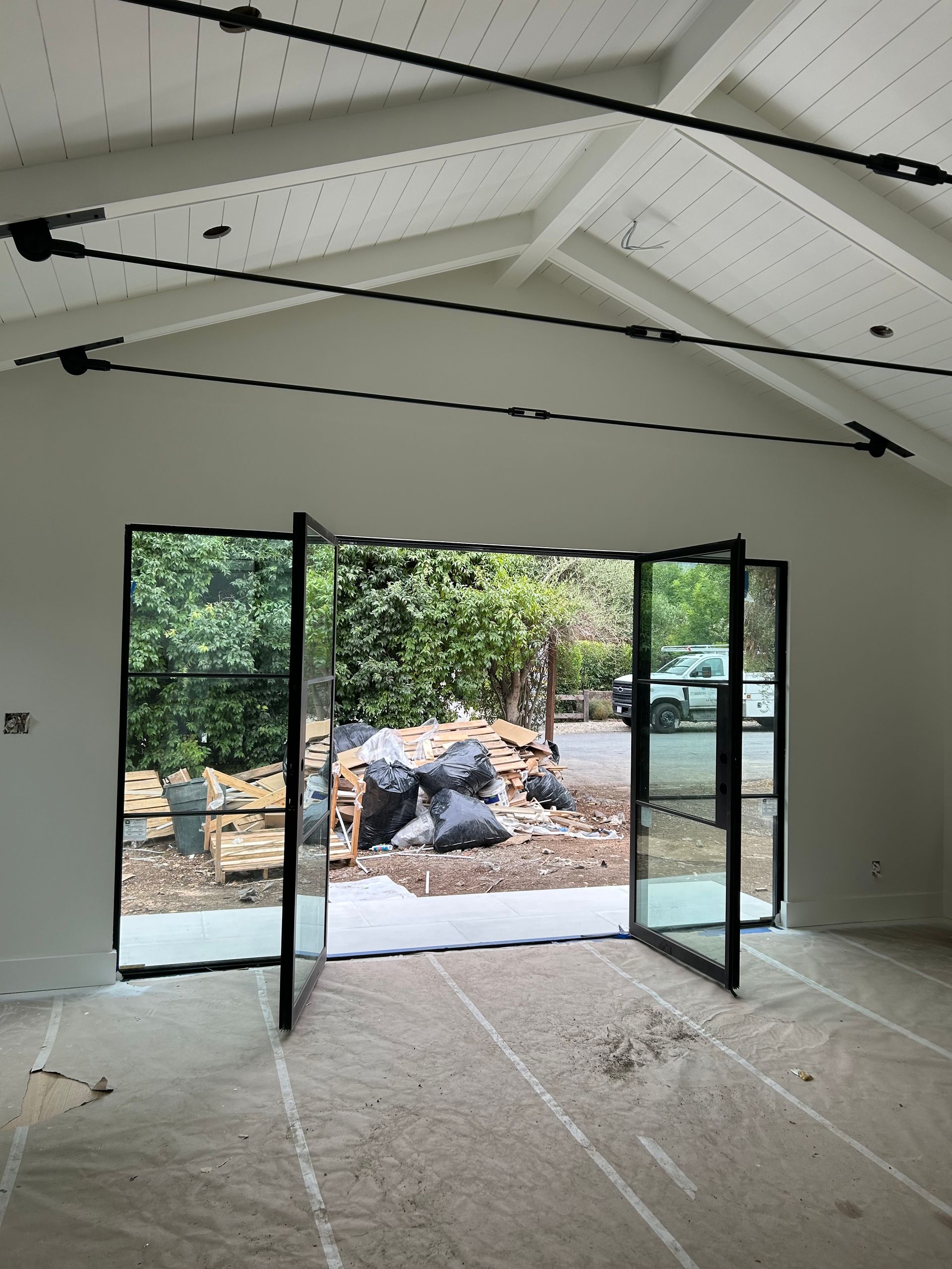 Interior with large black-framed doors open to a garden. White walls, wood ceiling beams, and concrete floor.