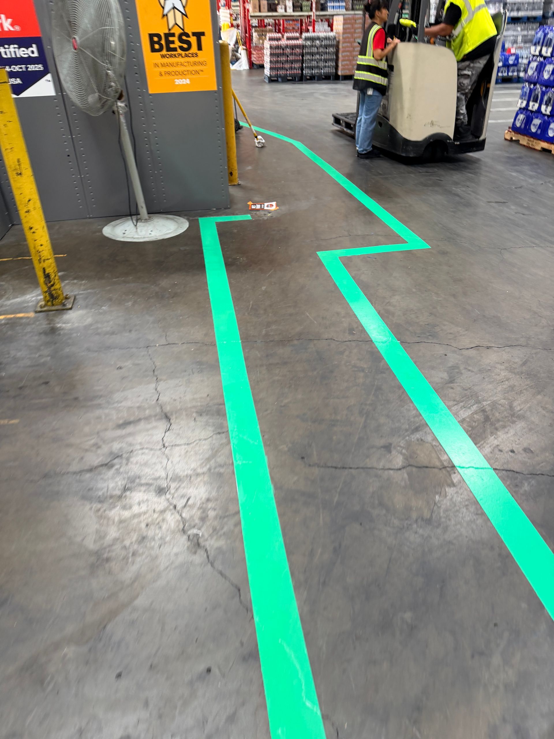 Green painted lines on a warehouse floor marking a path for forklifts. A worker guides a forklift.