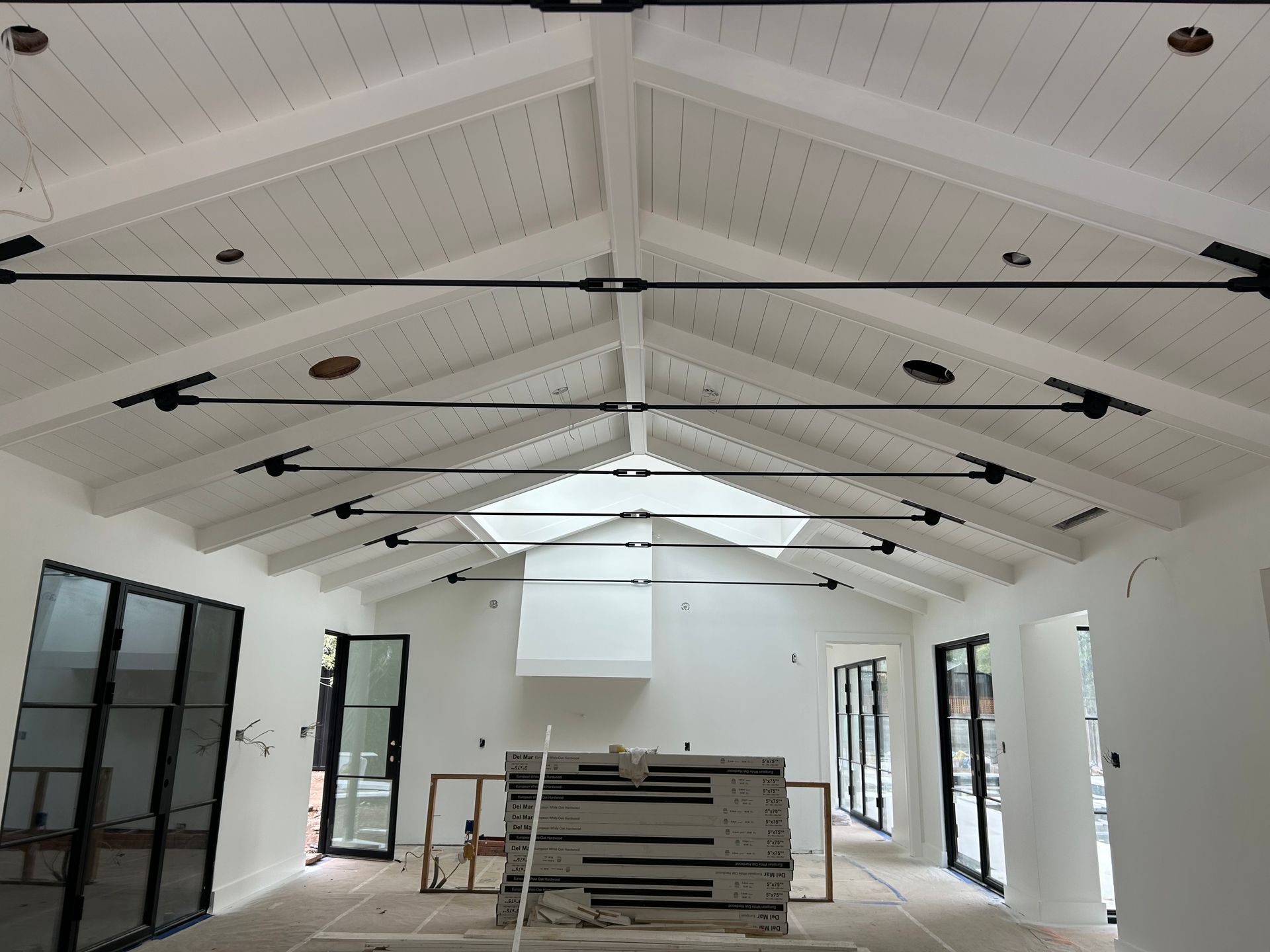 Interior view of a room under construction with white vaulted ceiling, black beams, and large windows.