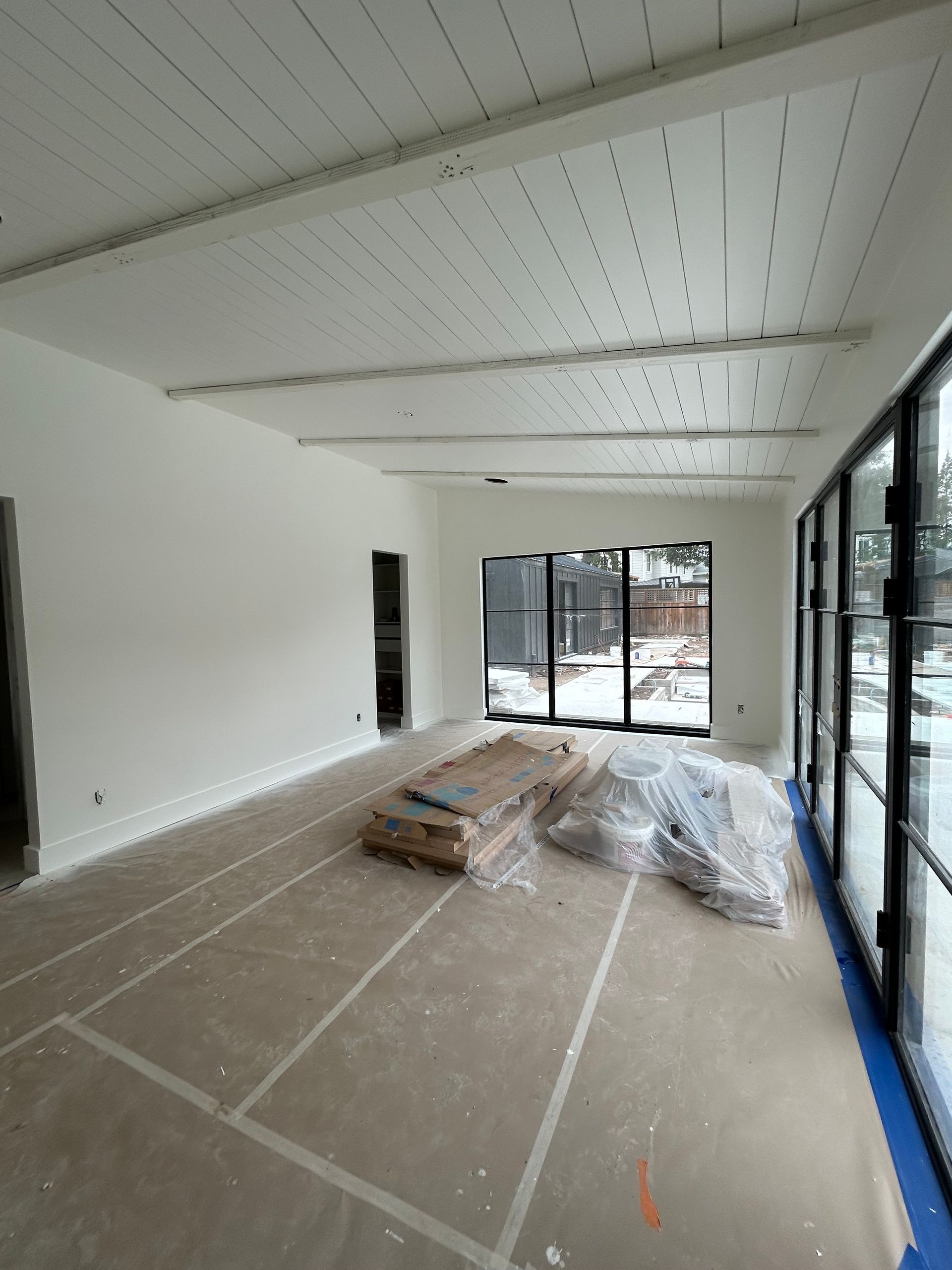 Empty room under construction with white walls, ceiling beams, and large windows.
