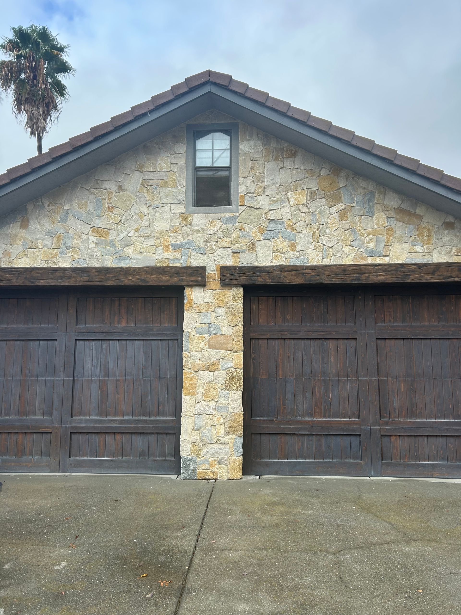 Two-car garage with brown doors, stone facade, and small window. A palm tree is visible in the background.