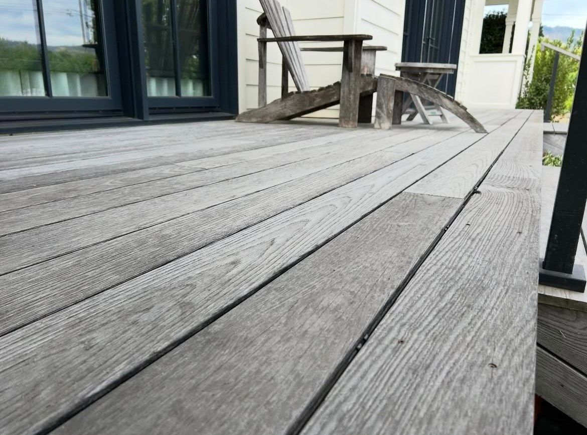 Weathered grey wooden deck with an Adirondack chair in the background, next to a building's sliding door.