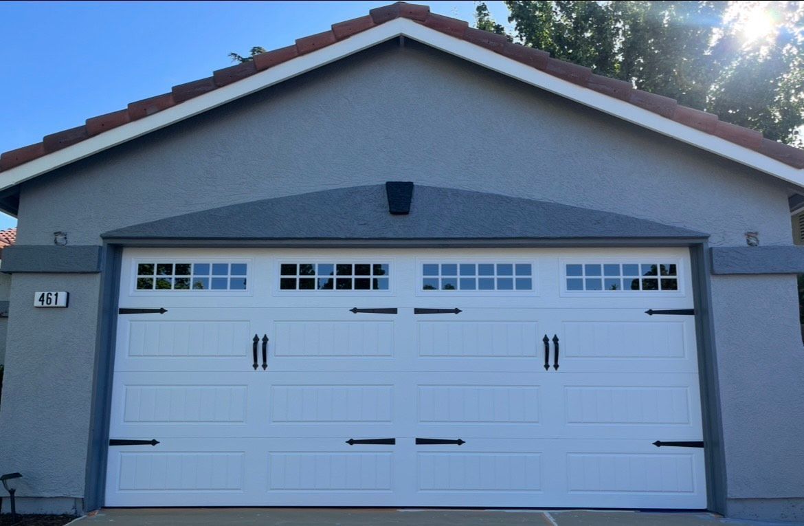 White garage door with decorative black hardware, framed by gray stucco and a red-tiled roof.