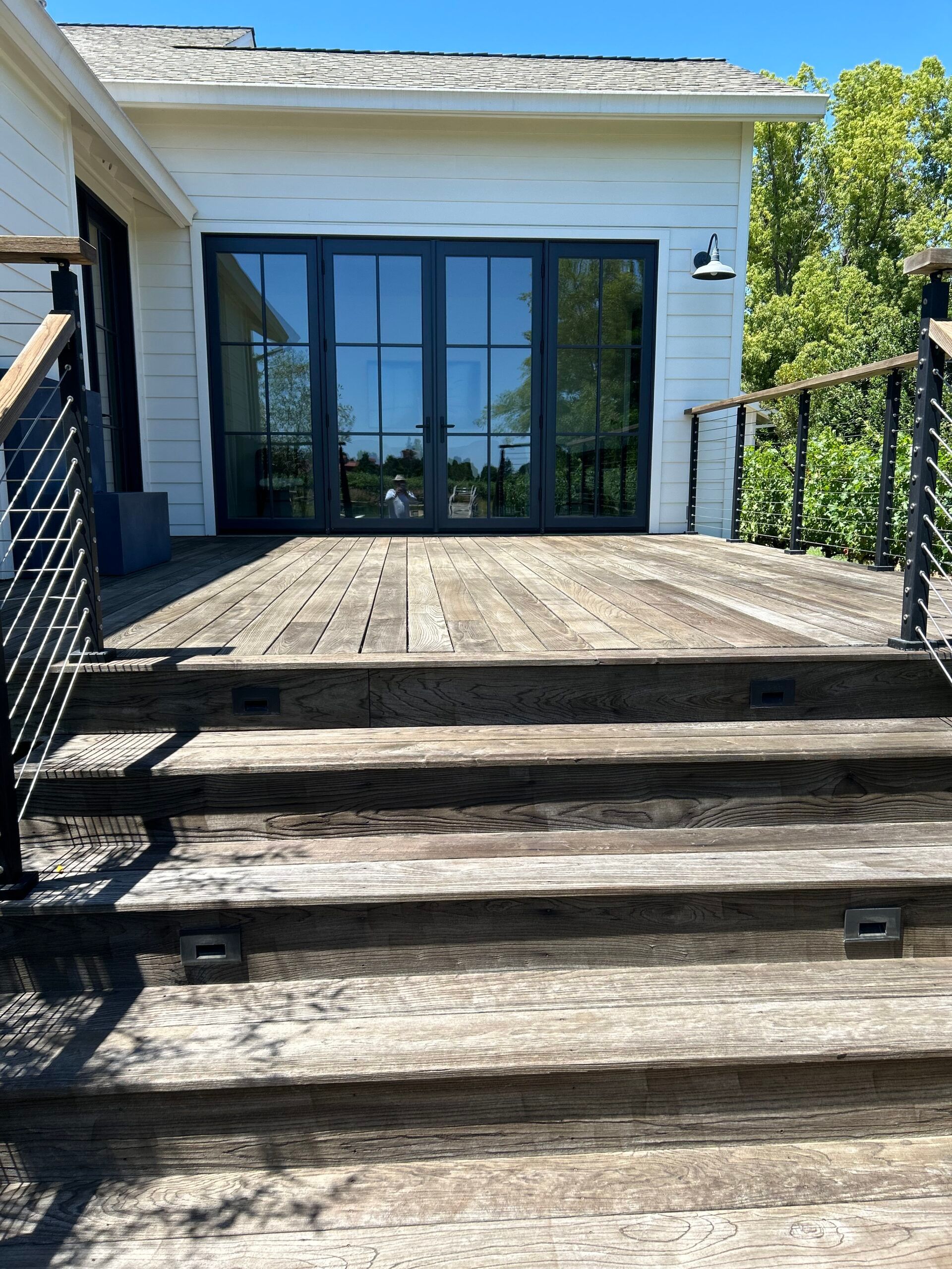 Wooden deck with stairs, black framed doors, and metal railings. Sunny day.