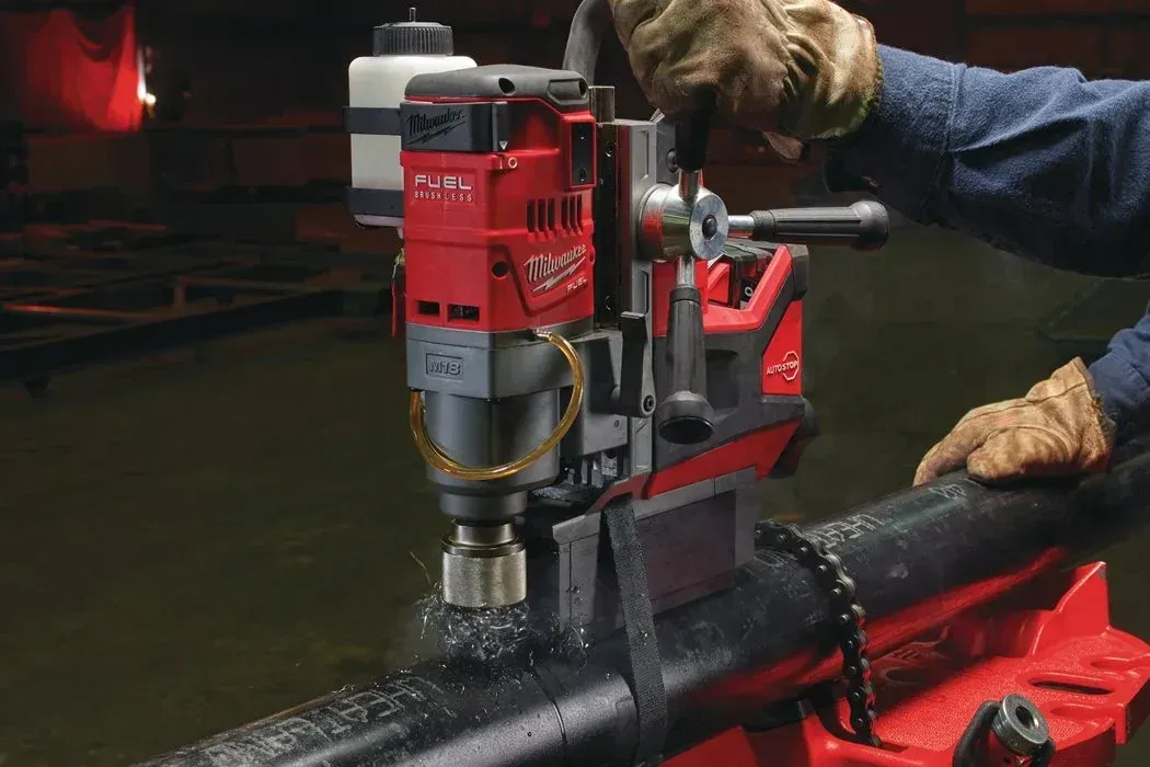 Una persona con indosso guanti da lavoro aziona un trapano a colonna magnetico Milwaukee di colore rosso, montato su un tubo orizzontale.