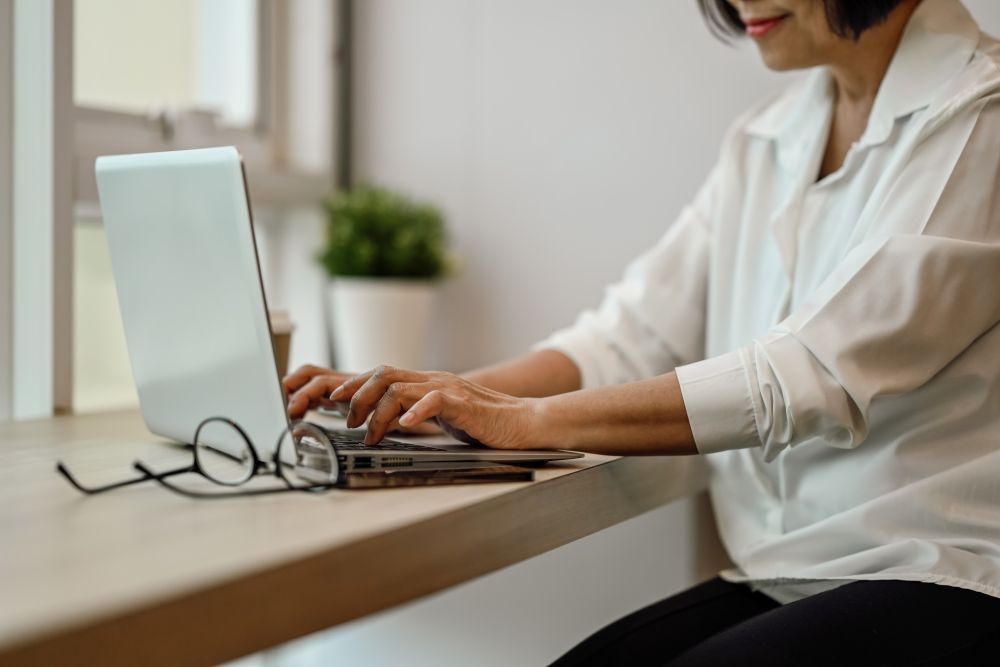 Person typing on a laptop at a desk beside glasses and a small white monitor in a bright workspace